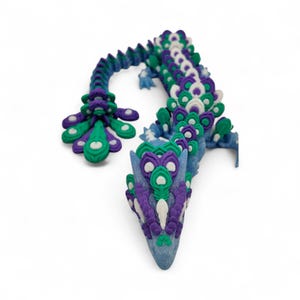 May include: A whimsical, handcrafted dragon sculpture in shades of blue, purple, green, and white. The dragon's body is adorned with intricate, feather-like details, and the head is pointed. The sculpture is set against a white background.