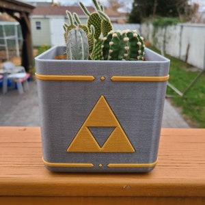 May include: A grey and gold 3D printed planter with a triangular design. The planter is filled with a variety of cacti.