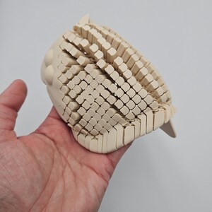 May include: A light-colored, wooden owl sculpture with a textured, geometric design. The owl's body is composed of numerous small, rectangular blocks, creating a unique visual effect. The sculpture is held in a hand against a plain background.