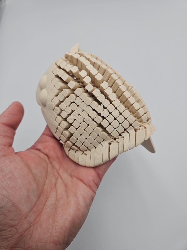 May include: A light-colored, wooden owl sculpture with a textured, geometric design. The owl's body is composed of numerous small, rectangular blocks, creating a unique visual effect. The sculpture is held in a hand against a plain background.