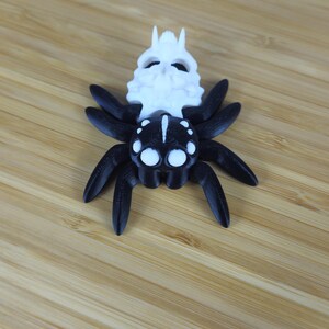 Puede incluir: Una ara&ntilde;a impresa en 3D en blanco y negro con una cabeza en forma de calavera. La ara&ntilde;a tiene ocho patas y grandes ojos blancos.