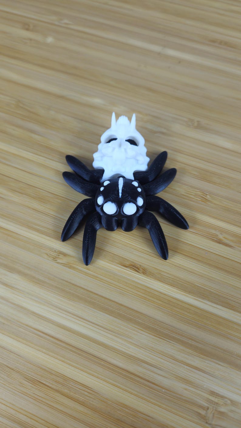 Puede incluir: Una ara&ntilde;a impresa en 3D en blanco y negro con una cabeza en forma de calavera. La ara&ntilde;a tiene ocho patas y grandes ojos blancos.