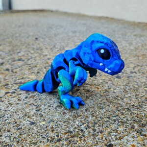 May include: A small, blue and green 3D printed dinosaur toy. The dinosaur has a segmented body and large, white eyes.