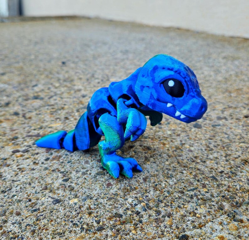 May include: A small, blue and green 3D printed dinosaur toy. The dinosaur has a segmented body and large, white eyes.