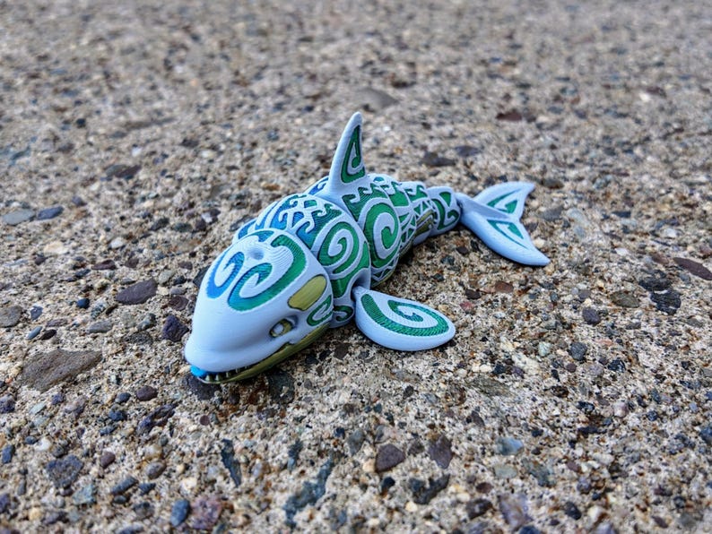May include: A blue and green toy shark with a swirling pattern. The shark has a toothy grin and is made of plastic.