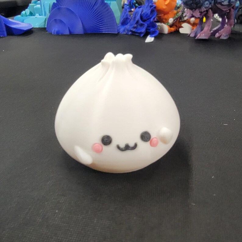 Peut inclure: Figurine de ravioli blanc imprim&eacute; en 3D avec un visage souriant, des yeux noirs et des joues roses. Le ravioli a un dessus froiss&eacute; et un petit bras. La figurine est sur une surface sombre.