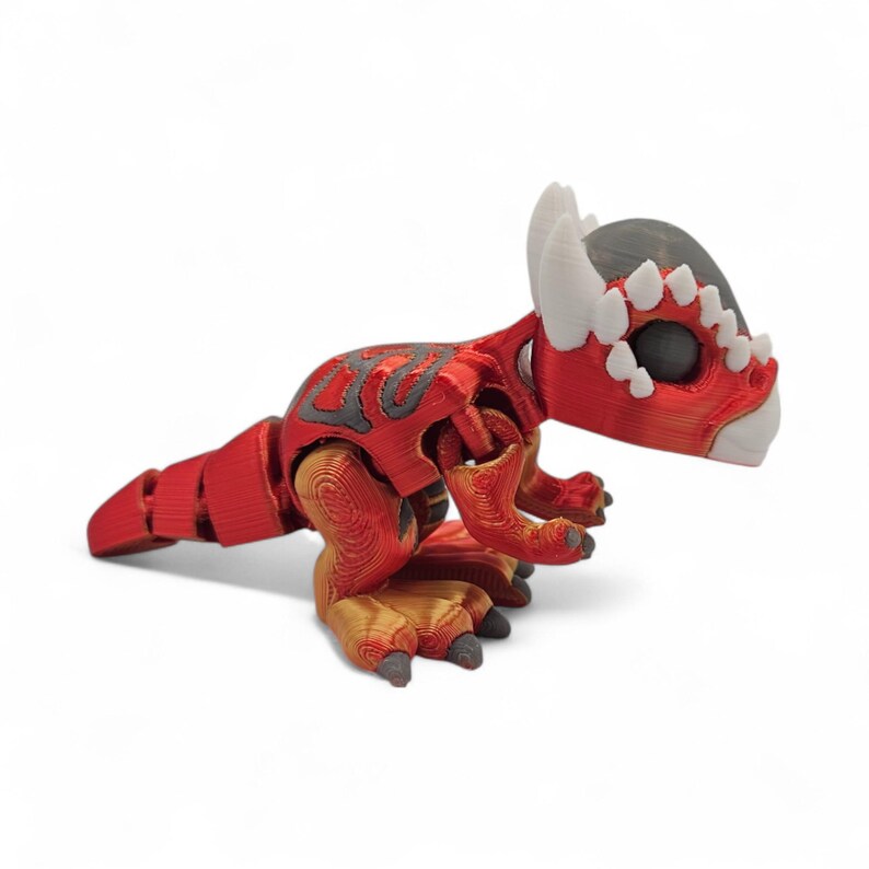 May include: A red and gray articulated dinosaur toy with a detailed design. The toy features a segmented body, with a head and tail. The dinosaur has white horns and teeth, and gold and gray accents on its feet and body.