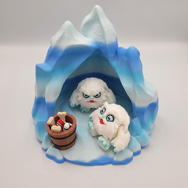 May include: A whimsical sculpture featuring two white, furry creatures with expressive faces, set within a blue and white iceberg-shaped structure. A small wooden barrel with bones and a red and white item sits nearby. The scene is playful and detailed.