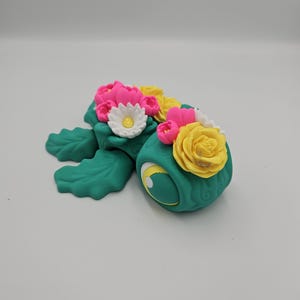 Pode incluir: Figura de tartaruga artesanal, na cor turquesa. &Eacute; adornada com um arranjo floral de flores rosa, amarelas e brancas. O olho da tartaruga &eacute; amarelo com uma pupila preta. A figura est&aacute; sobre um fundo branco liso.