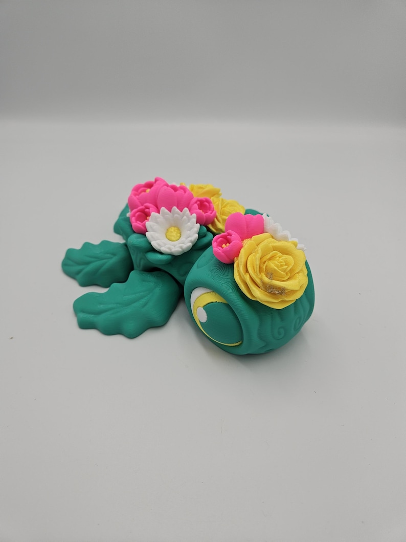 Pode incluir: Figura de tartaruga artesanal, na cor turquesa. &Eacute; adornada com um arranjo floral de flores rosa, amarelas e brancas. O olho da tartaruga &eacute; amarelo com uma pupila preta. A figura est&aacute; sobre um fundo branco liso.