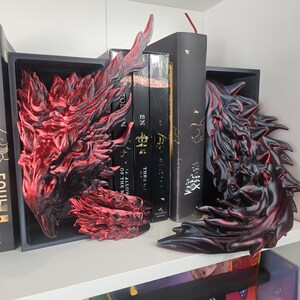 May include: A set of dragon-themed bookends in red and black. The bookends have a detailed, textured design and are placed on a white shelf with books. The dragon wings are the main focus, adding a fantasy element to the bookshelf.