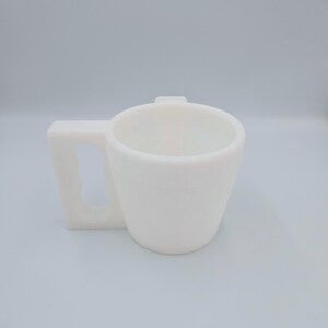 Pu&ograve; includere: Una tazza in ceramica bianca con manico quadrato. La tazza ha un corpo cilindrico che si assottiglia leggermente verso la base. Il manico ha una forma rettangolare solida con una piccola linguetta rialzata sul bordo superiore della tazza.