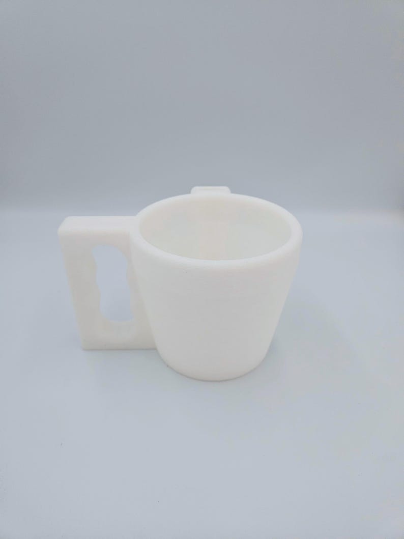 Pu&ograve; includere: Una tazza in ceramica bianca con manico quadrato. La tazza ha un corpo cilindrico che si assottiglia leggermente verso la base. Il manico ha una forma rettangolare solida con una piccola linguetta rialzata sul bordo superiore della tazza.
