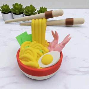 K&ouml;nnte beinhalten: Ein farbenfroher Ramen-Bowl mit gelben Nudeln, rosa Garnelen, einem gekochten Ei und gr&uuml;nem Seetang. Beige Essst&auml;bchen mit braunen B&auml;ndern sind &uuml;ber der Sch&uuml;ssel positioniert. Die Sch&uuml;ssel ist rot mit wei&szlig;em Boden. Das Bild deutet auf einen dekorativen Artikel hin.