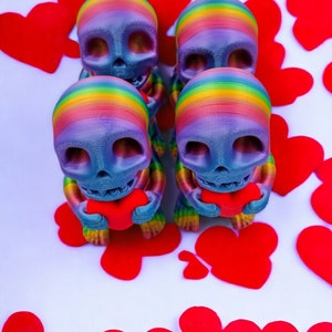 May include: Four rainbow-coloured skull figurines holding red hearts. The figurines are arranged in a row, with the hearts facing forward. The background is white with red hearts scattered around the figurines.