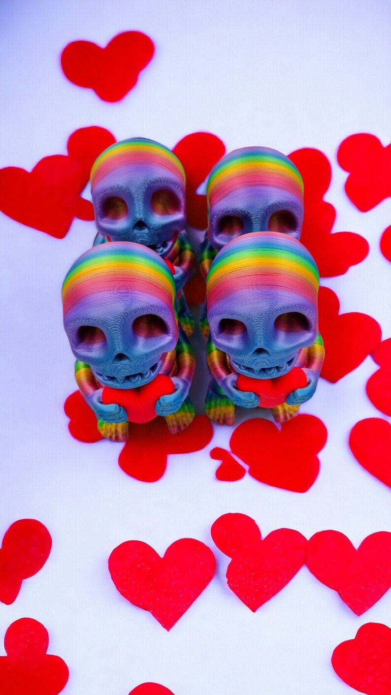 May include: Four rainbow-coloured skull figurines holding red hearts. The figurines are arranged in a row, with the hearts facing forward. The background is white with red hearts scattered around the figurines.