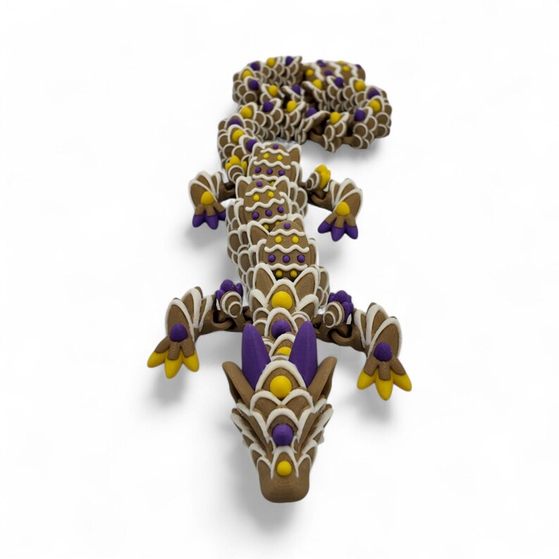 May include: A brown dragon figurine with white, yellow, and purple accents. The dragon is decorated with intricate patterns and details, and is lying on a white surface. The dragon's head is facing the viewer.