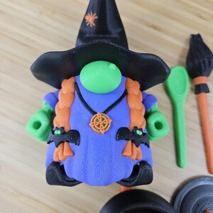 Puede incluir: Una caprichosa figura de gnomo bruja de Halloween con un sombrero negro, pelo naranja y un vestido azul. El gnomo tiene manos verdes y una nariz verde. Los accesorios incluyen un caldero, una cuchara y una escoba. El sombrero tiene una estrella naranja.