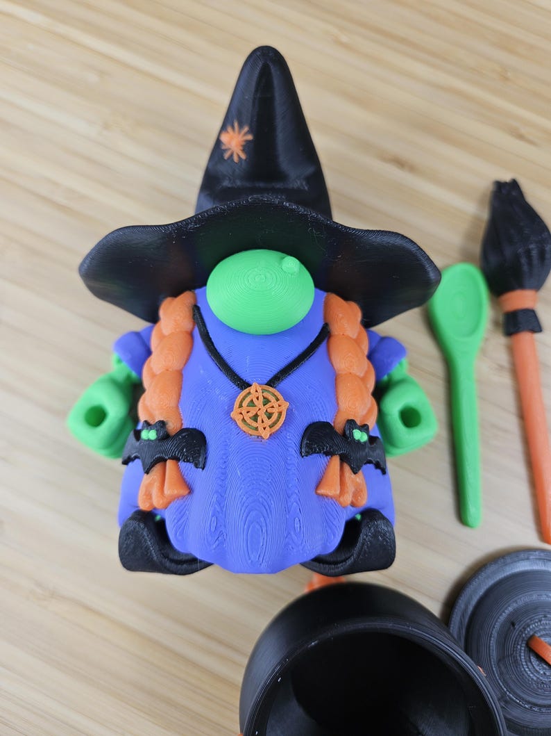 Puede incluir: Una caprichosa figura de gnomo bruja de Halloween con un sombrero negro, pelo naranja y un vestido azul. El gnomo tiene manos verdes y una nariz verde. Los accesorios incluyen un caldero, una cuchara y una escoba. El sombrero tiene una estrella naranja.