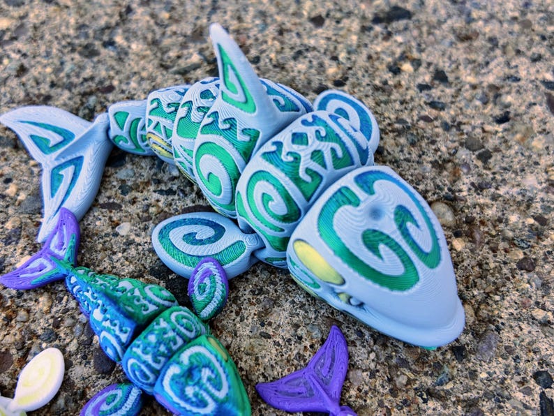 May include: A blue and green 3D printed shark with a swirling pattern. The shark is made of plastic and has a detailed design.