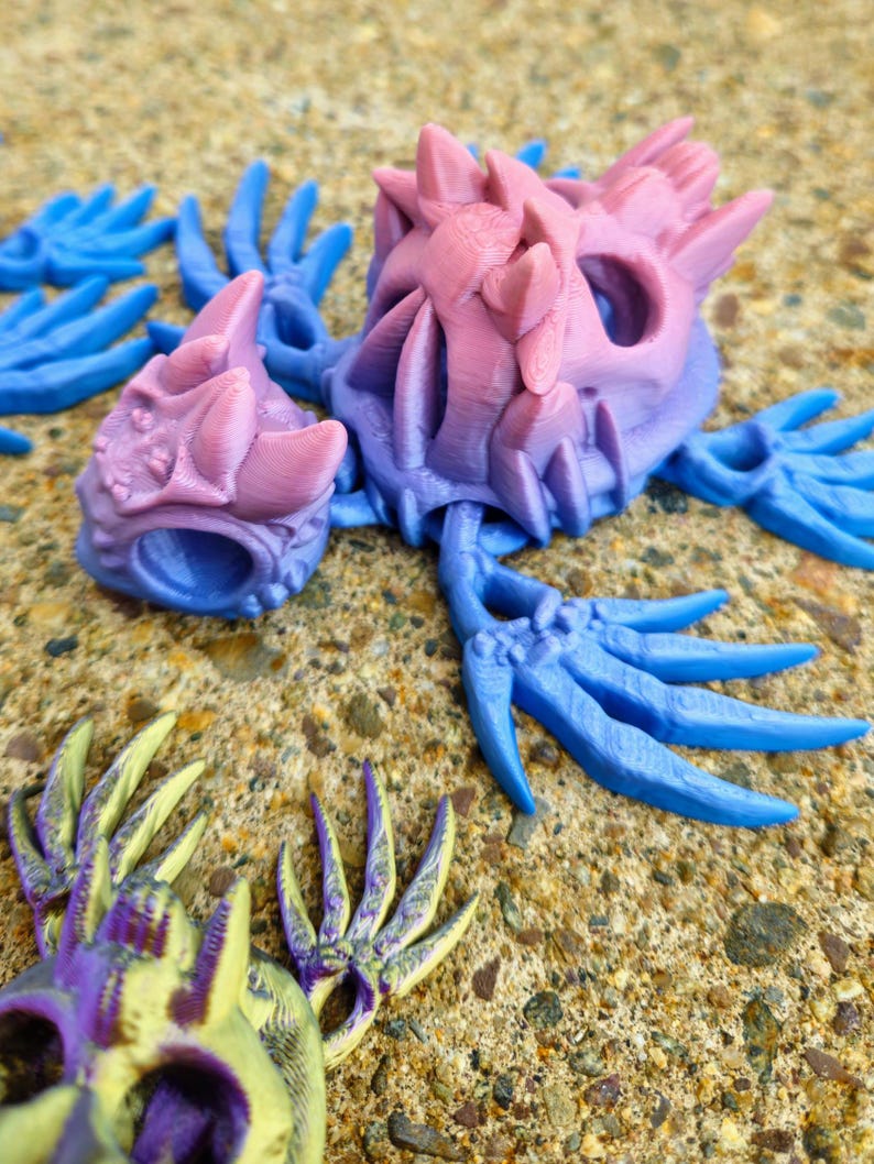 Peut inclure: Un ensemble de sculptures de mains squelettiques imprim&eacute;es en 3D en bleu, rose et jaune avec des accents violets. Les mains sont pos&eacute;es dans diff&eacute;rentes positions, certains doigts &eacute;tant tendus et d'autres recourb&eacute;s.
