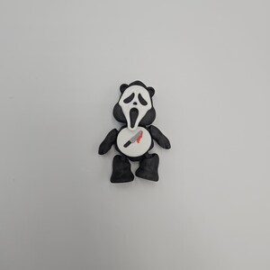 May include: A black and white figurine resembling a panda, with a Scream mask face. The figurine has black arms, legs, and ears. A white circle on the chest features a knife with a red blood stain.