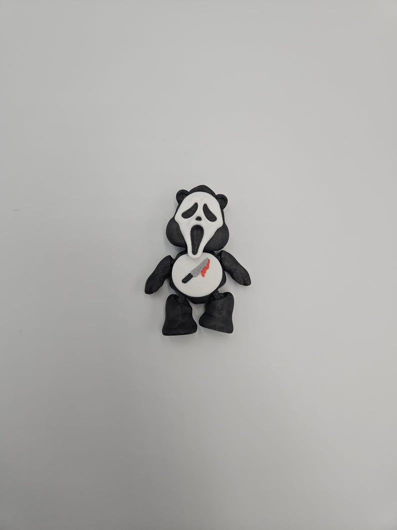 May include: A black and white figurine resembling a panda, with a Scream mask face. The figurine has black arms, legs, and ears. A white circle on the chest features a knife with a red blood stain.