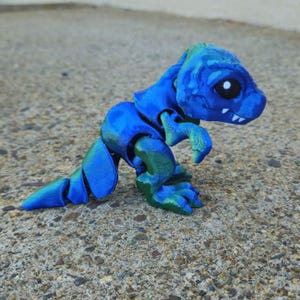 May include: A small, blue and green 3D printed dinosaur toy. The dinosaur is standing on all fours and has a large head with a wide mouth and big eyes.