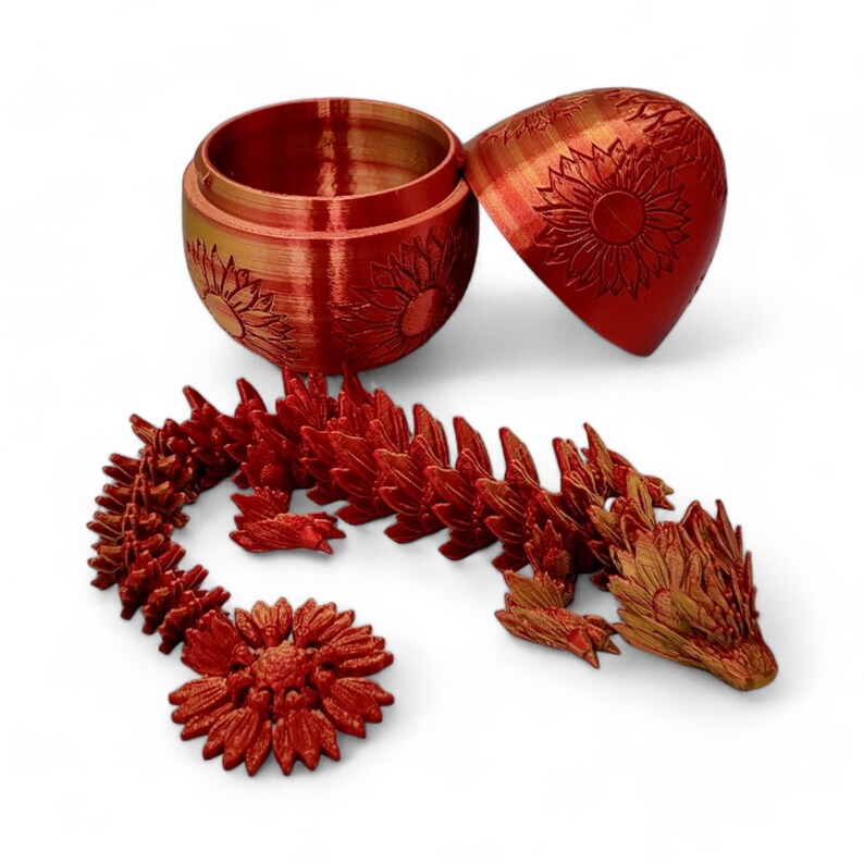 Puede incluir: Una figurita de drag&oacute;n roja impresa en 3D con detalles de girasol. El drag&oacute;n est&aacute; enroscado y tiene la boca cerrada. El drag&oacute;n est&aacute; al lado de una caja roja en forma de huevo impresa en 3D con un patr&oacute;n de girasol y la tapa abierta.
