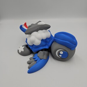 Pode incluir: Uma figura de personagem de desenho animado azul, cinza e branco. A figura tem um chap&eacute;u, asas e p&eacute;s azuis. O personagem tem um peito branco e fofo e uma cabe&ccedil;a cinza com um detalhe branco e vermelho.