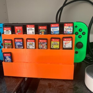 May include: An orange plastic stand holds 18 Nintendo Switch game cartridges. A blue and green Nintendo Switch console is on the right side of the stand.