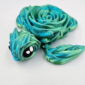 May include: A green and blue 3D printed rose with a whimsical, cartoon-like face. The rose is made of layered, textured plastic.