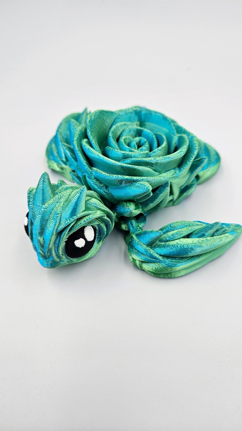 May include: A green and blue 3D printed rose with a whimsical, cartoon-like face. The rose is made of layered, textured plastic.