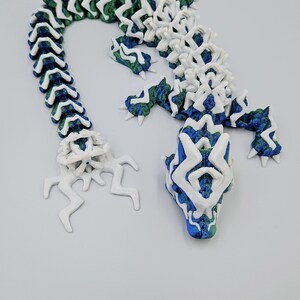May include: A 3D printed lizard figurine with a white and green body and a blue head. The lizard is made up of interlocking pieces that form a segmented body.