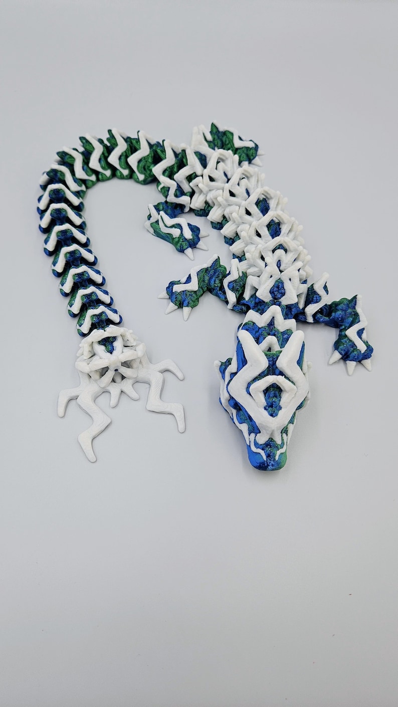 May include: A 3D printed lizard figurine with a white and green body and a blue head. The lizard is made up of interlocking pieces that form a segmented body.