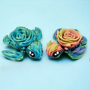 May include: Two handmade polymer clay turtle figurines with rose-shaped shells. One turtle has a blue and green shell with a black eye, and the other has a rainbow-colored shell with a black eye.