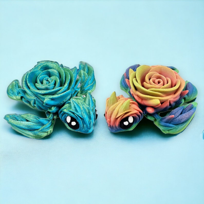 May include: Two handmade polymer clay turtle figurines with rose-shaped shells. One turtle has a blue and green shell with a black eye, and the other has a rainbow-colored shell with a black eye.