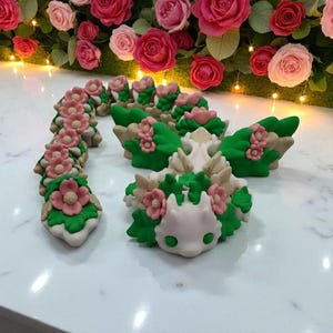 May include: A collection of handcrafted, decorative figurines. The figurines feature a white base with green, pink, and beige floral and leaf accents. The background includes a display of pink and red roses.