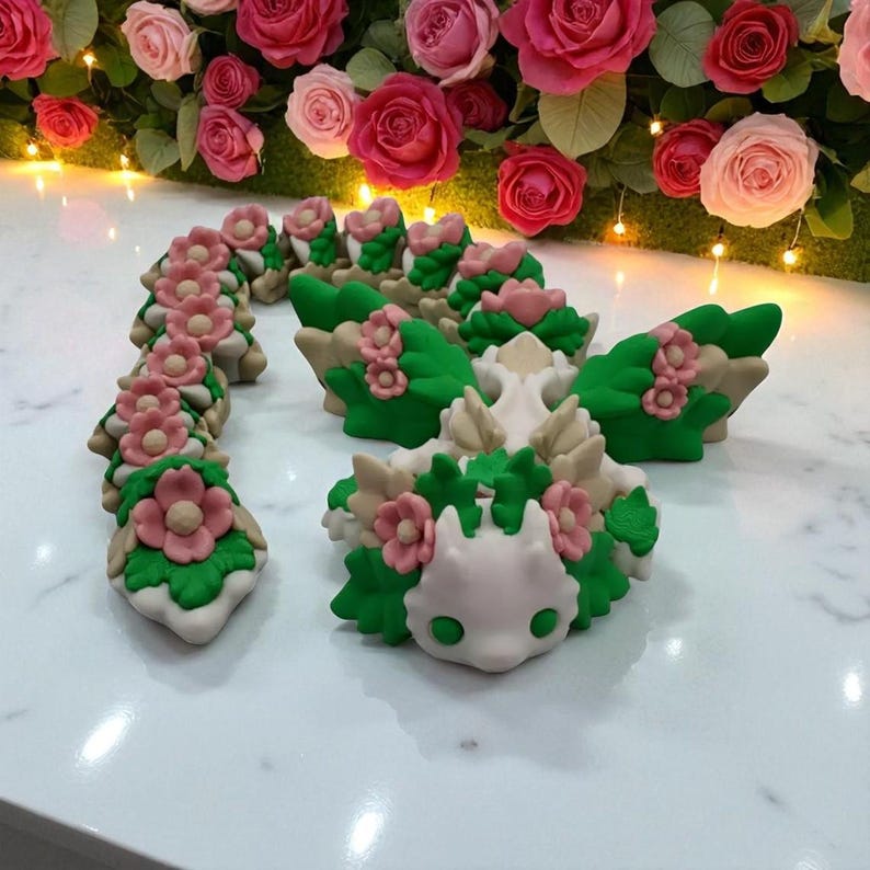 May include: A collection of handcrafted, decorative figurines. The figurines feature a white base with green, pink, and beige floral and leaf accents. The background includes a display of pink and red roses.