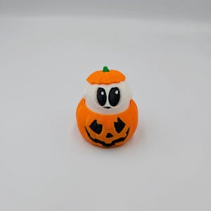 May include: A small, white ghost figurine with black eyes is inside a miniature orange pumpkin with a carved jack-o'-lantern face. The pumpkin has a green stem.