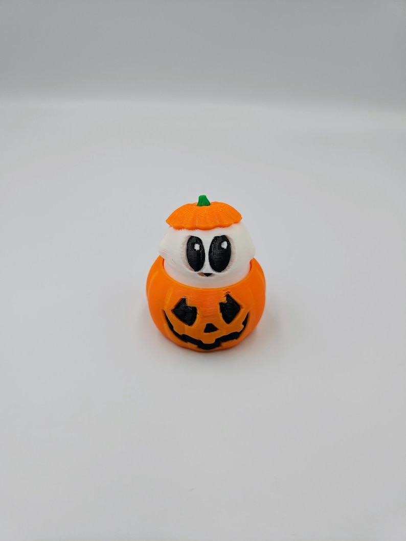 May include: A small, white ghost figurine with black eyes is inside a miniature orange pumpkin with a carved jack-o'-lantern face. The pumpkin has a green stem.
