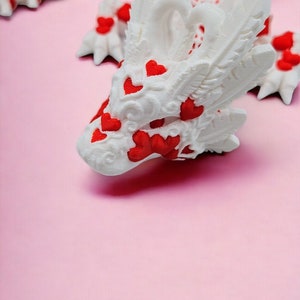 Op de afbeelding: Witte drakenfiguur met rode harten op een roze achtergrond. De draak heeft ingewikkelde details en een speels ontwerp.