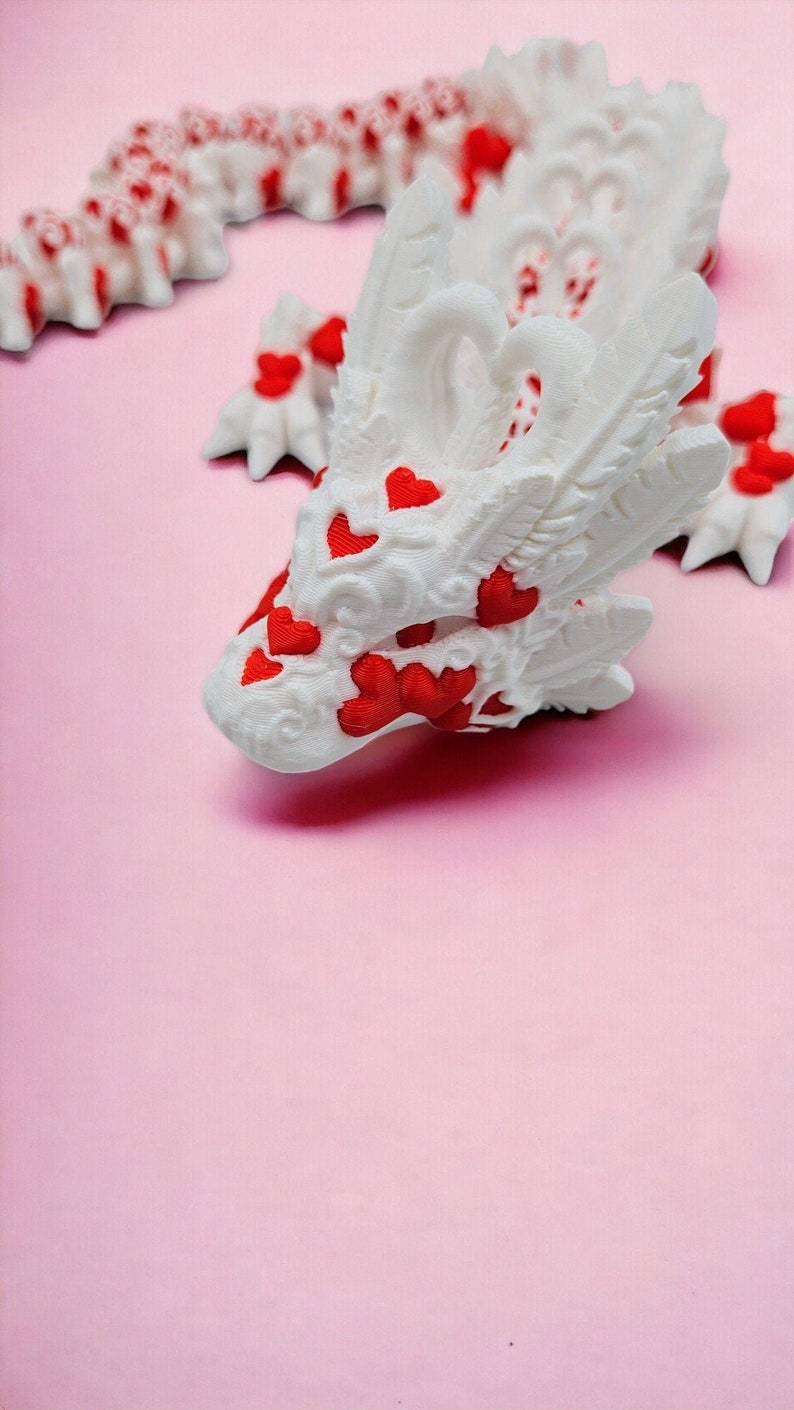Op de afbeelding: Witte drakenfiguur met rode harten op een roze achtergrond. De draak heeft ingewikkelde details en een speels ontwerp.