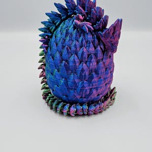 May include: A 3D printed dragon egg figurine with iridescent blue, purple, and green scales. The egg is curled up with its tail wrapped around its body.