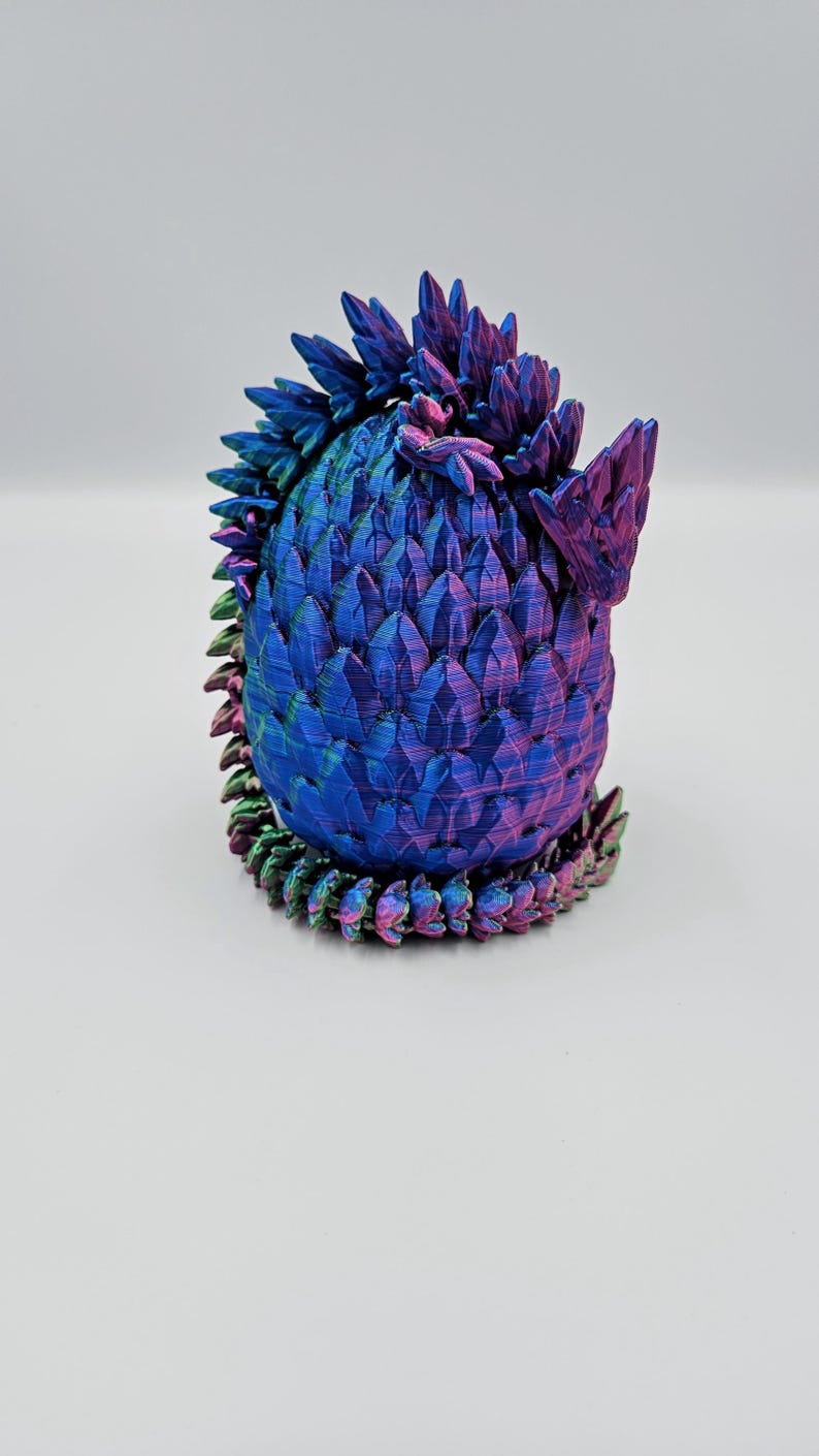 May include: A 3D printed dragon egg figurine with iridescent blue, purple, and green scales. The egg is curled up with its tail wrapped around its body.