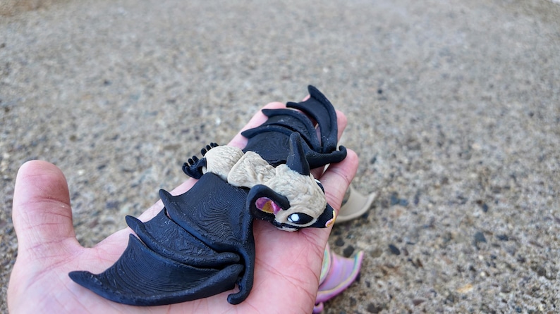 May include: A black and beige bat figurine with wings spread out. The bat has a detailed texture and a pink ear.