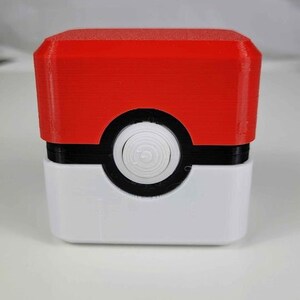 May include: A red, white, and black 3D printed box with a round button on the front. The box is shaped like a Pokeball.