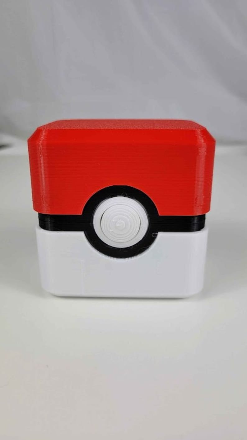 May include: A red, white, and black 3D printed box with a round button on the front. The box is shaped like a Pokeball.