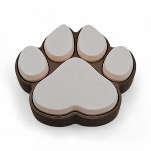 May include: A brown and white paw-shaped item. The item is a paw print design with five pads. The pads are white and the surrounding border is brown. The item is likely a coaster or decorative piece.