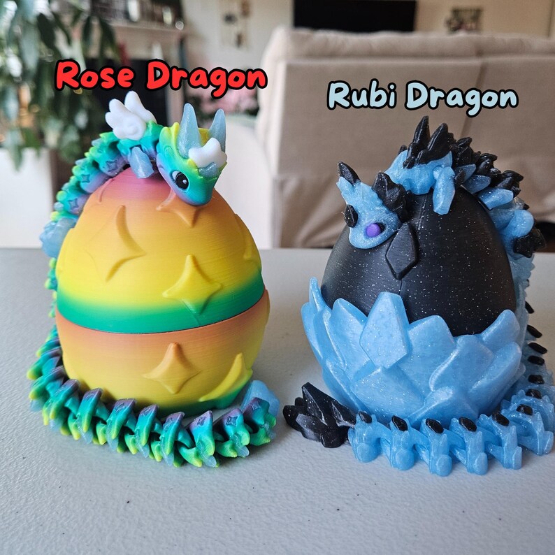 May include: Two 3D printed dragon figurines, one with a rainbow egg and the other with a blue and black egg. The dragon with the rainbow egg is labeled "Rose Dragon" and the dragon with the blue and black egg is labeled "Rubi Dragon".
